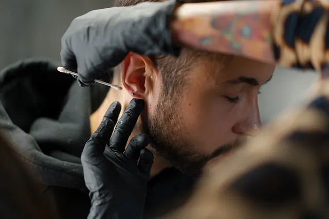 Ein Mann bekommt ein Piercing am Ohr gestochen. Die Piercing Pflege erfolgt mit einem Wattestäbchen. 